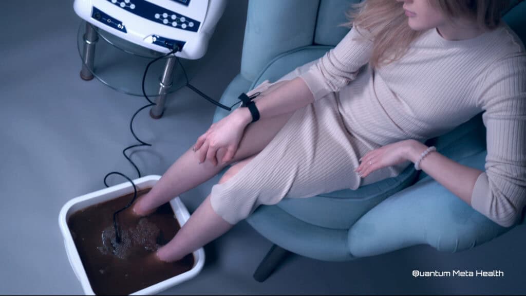 Person using a Quantum Meta Health ionic detox machine with their feet in a basin of water, connected to electrodes, undergoing a detoxification process.