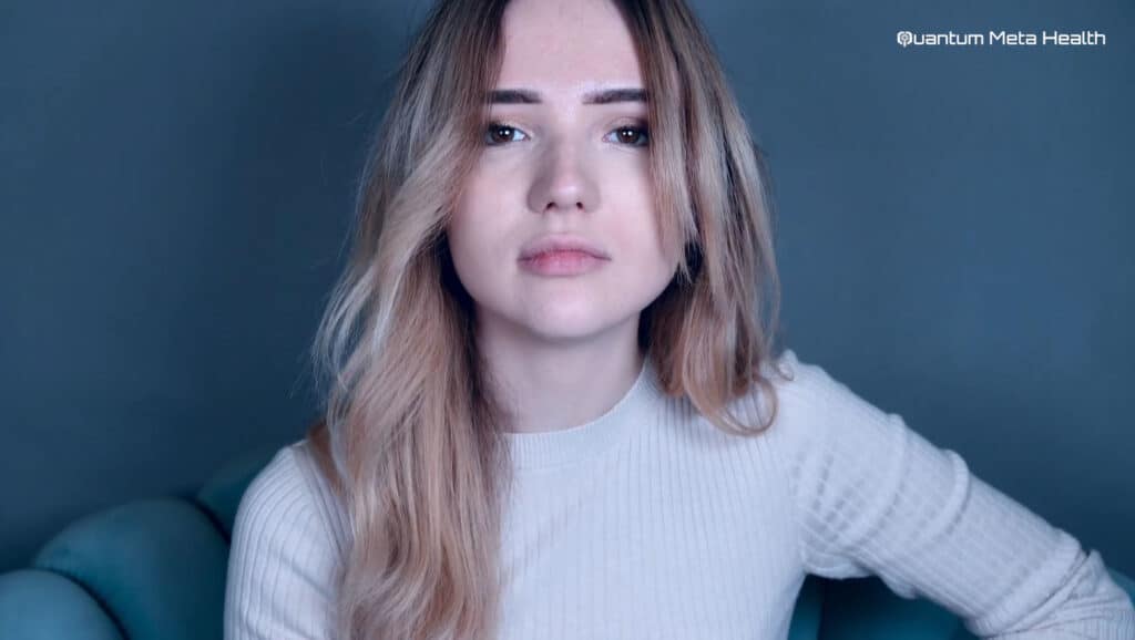 Close-up of a woman during a Quantum Meta Health ionic detoxification session. She is sitting in a chair, looking directly at the camera with a calm expression. The focus is on her upper body and face, highlighting her relaxed state. The Quantum Meta Health logo is visible in the background, emphasizing the context of the session. The image represents a serene and therapeutic environment, showcasing the benefits of the ionic detox process provided by Quantum Meta Health.