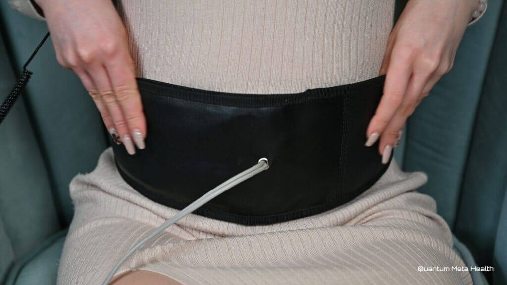 Close-up of a woman undergoing an ionic detoxification session using a Quantum Meta Health heated waist belt. The woman is comfortably seated, wearing a light-colored ribbed dress. The detox machine's electrodes are connected to the waist belt, facilitating the detox process. The Quantum Meta Health logo is visible, reinforcing the brand's advanced detox technology. This image emphasizes the therapeutic benefits and health-focused approach of the ionic detox session, showcasing the equipment's effectiveness in promoting wellness and detoxification.