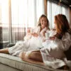 Two women in white bathrobes enjoying tea in a serene, sunlit wellness setting representing the Quantum Meta Health holistic experience